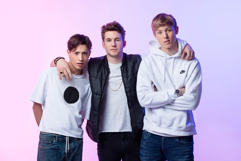Tre unge gutter i hvite og sorte t-skjorter og hettegensere står tett med hånden rundt skulderen på hverandre foran en dus rosa bakgrunn. 
