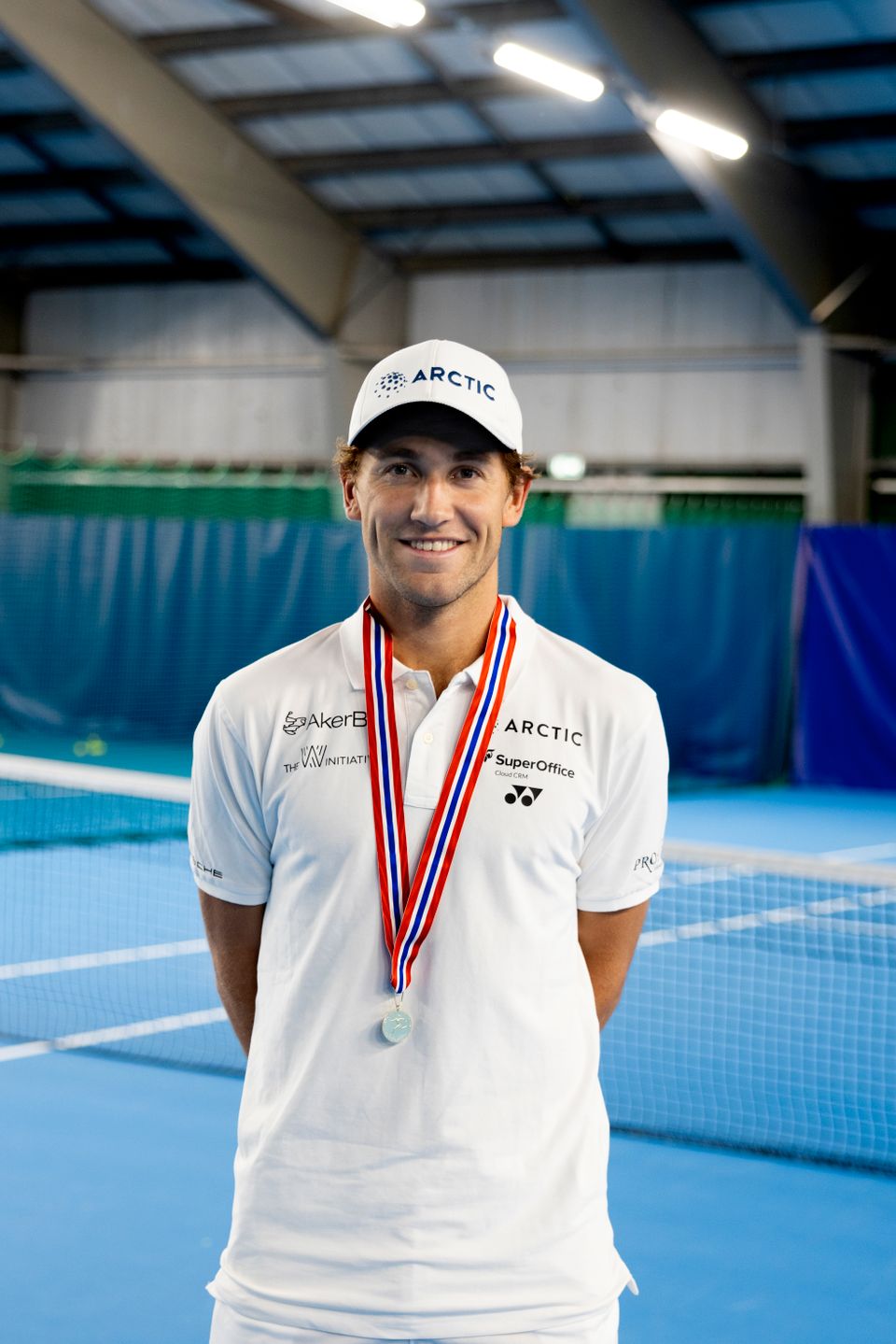 En mann med hvit caps, hvit t-skjorte og en medalje rundt halsen smiler.
