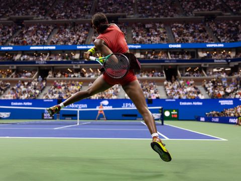 En kvinnelig tennisspiller ses bakfra mens hun slenger racketen rundt kroppen og hopper opp i luften, med beina spredt, på en hardcourt-bane med fulle tribuner.