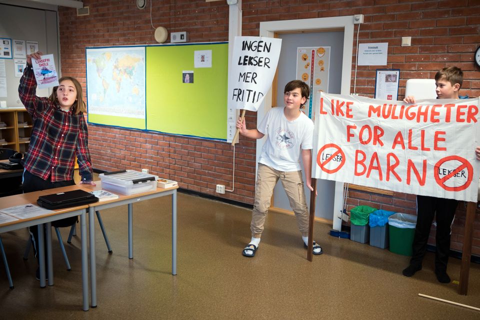 En jente med rød-rutete skjorte holder opp en pamflett og to gutter holder en fane med rød skrift på inne i et klasserom.