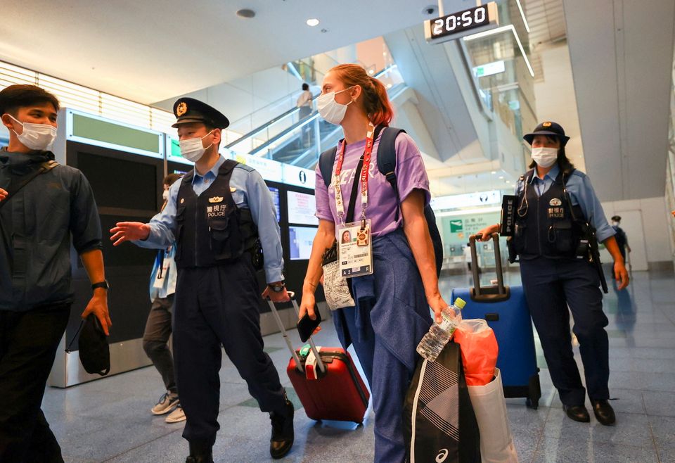 OL-utøveren triller på en koffert, omringet av vakter på flyplassen i Tokyo. 