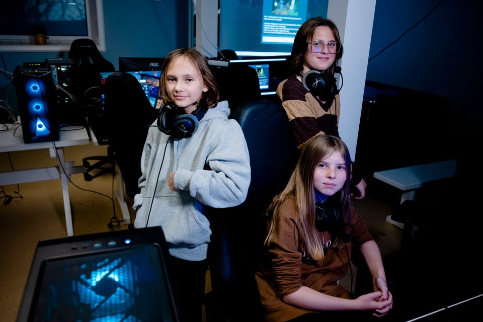 Tre jenter sitter og står ved en PC i et rom med mange PCer og de har store høretelefoner rundt halsen. 