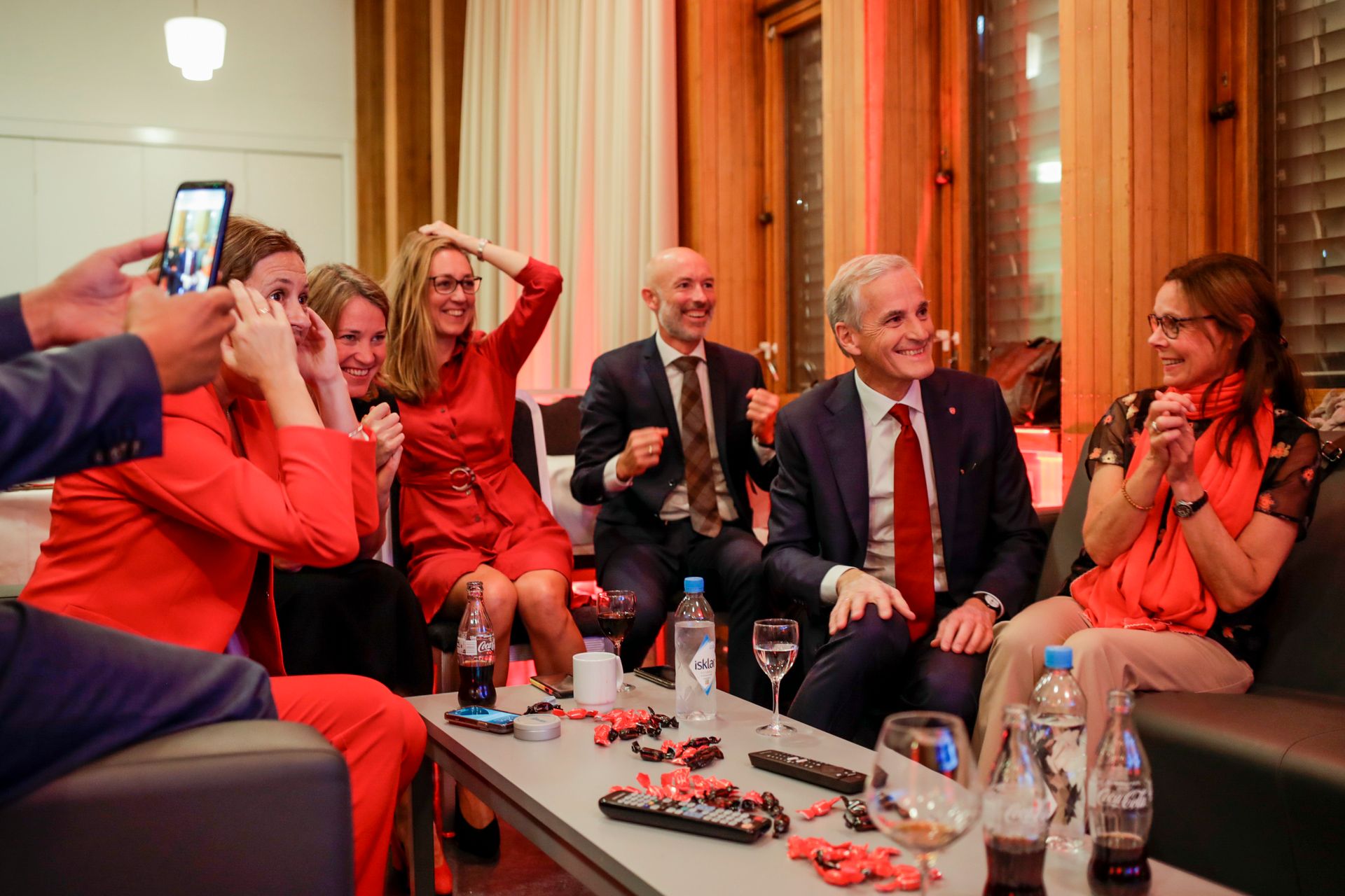 Støre, med rødt slips, og en gruppe støttespillere sitter rundt et bord og smiler og jubler, mens de antakelig ser mot en skjerm eller person som deler foreløpige resultater fra opptellingen.