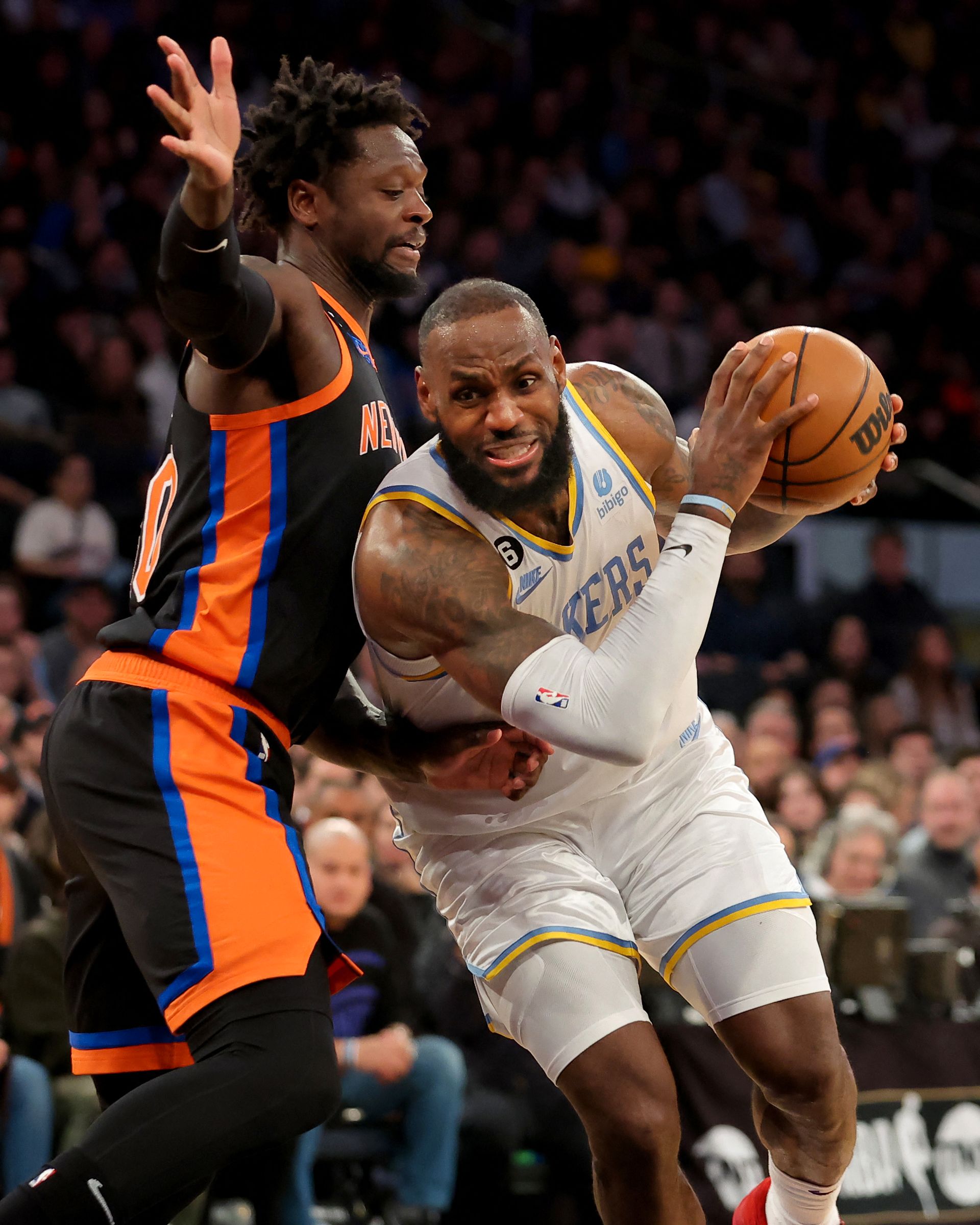 En mann med kort hår og hvit drakt som det står "Lakers" på, holder en basketball og løper forbi en annen spiller som prøver å stanse ham.