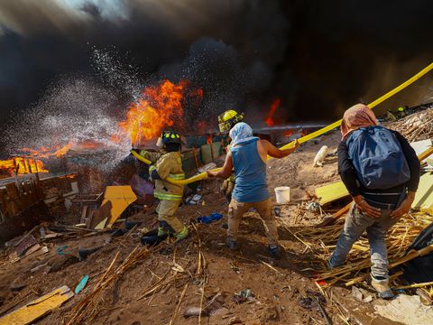 En brannmann i gule klær og to mennesker i sivile klær holder en gul brannslange og spyler vann mot store, oransje flammer og tykk, svart røyk. 