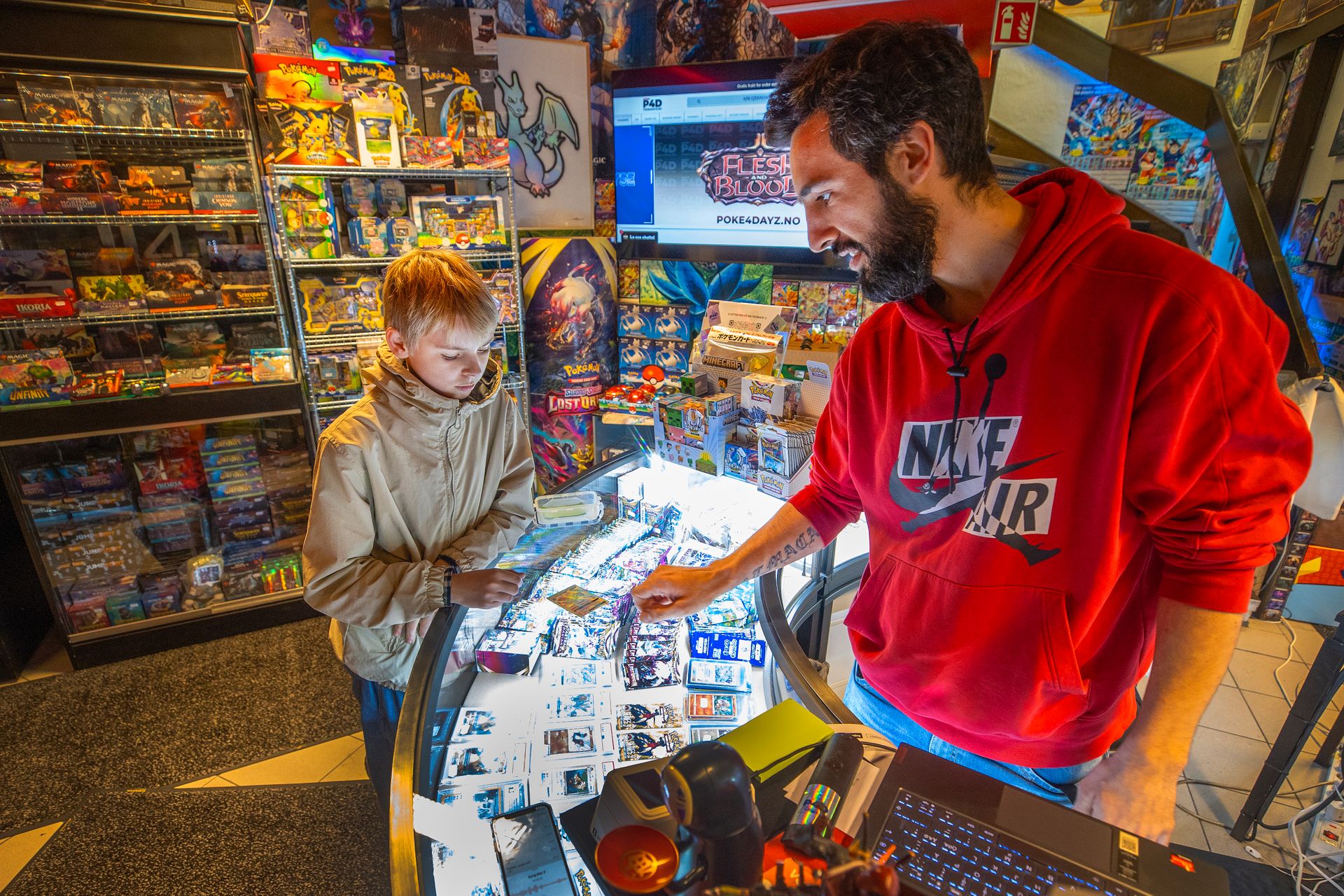 En gutt med lyst hår står ved disken inne i en butikk med veldig mange figurer, lys og kort, og bak disken står en mann i rød hettegenser som ser på kortet som gutten har lagt på disken