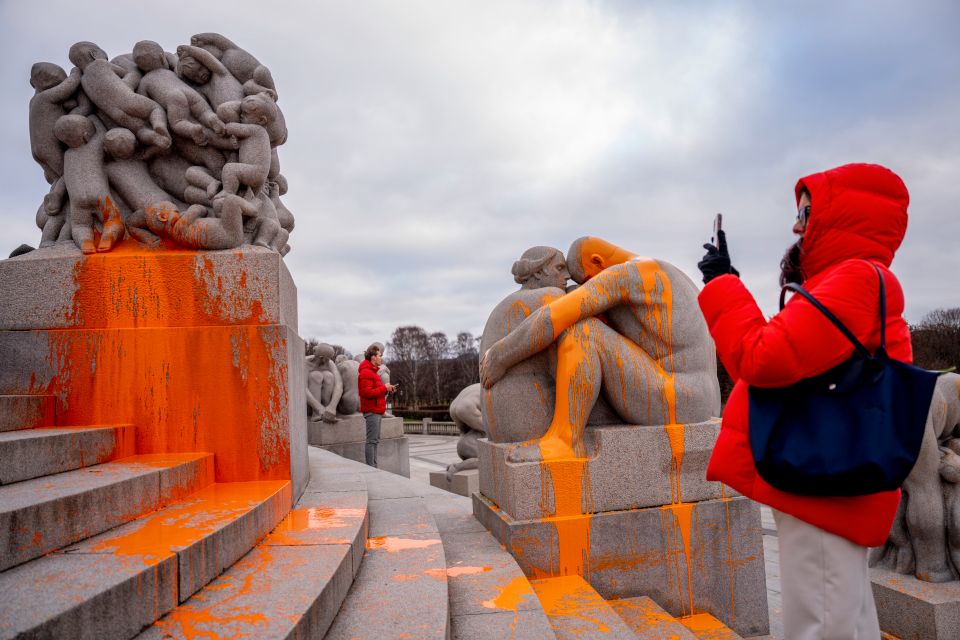 En person står og tar bilde av statuer av stein som det er kastet oransje maling på.