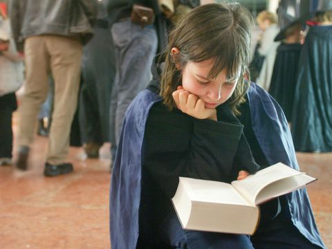 En jente med kappe sitter ned og leser en tykk Harry Potter-bok mens det er mange mennesker i bakgrunnen