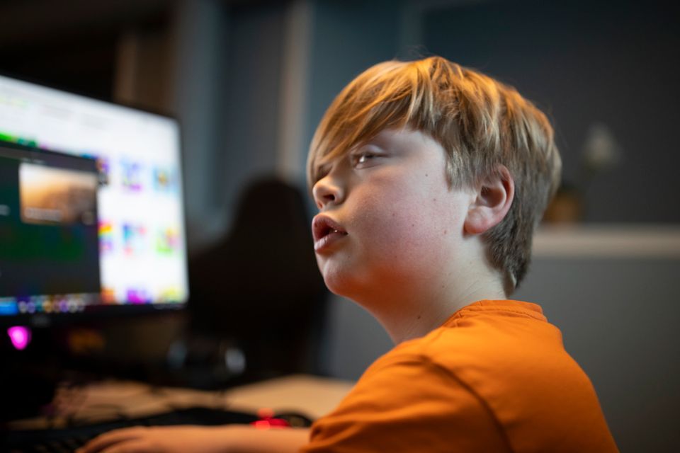 En gutt med lyst hår og oransje t-skjorte som sitter ved en PC-skjerm. 