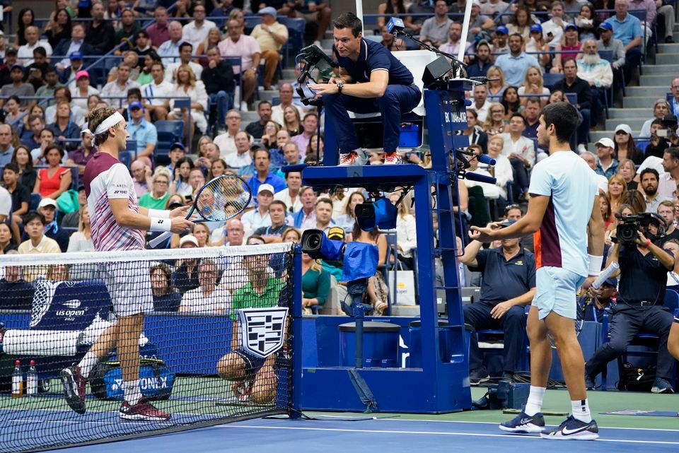To tennisspillere, på hver sin side av nettet, går mot dommeren, som sitter på et høyt, blått podium, mens mange publikummere sitter på tribunen. 