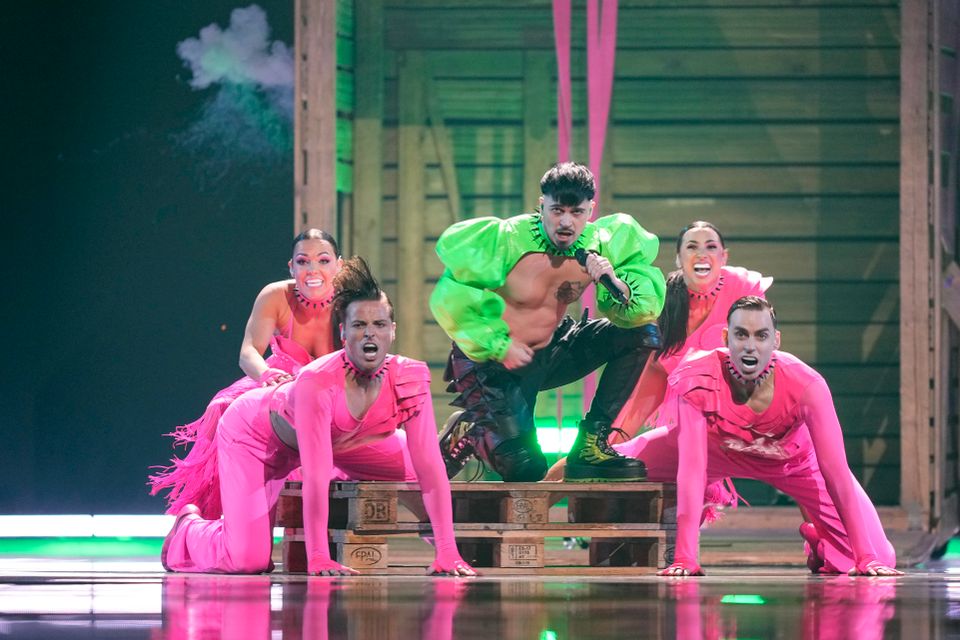 Fem personer på en scene, én med grønne klær og mikrofon, og fire som danser foran med rosa klær.
