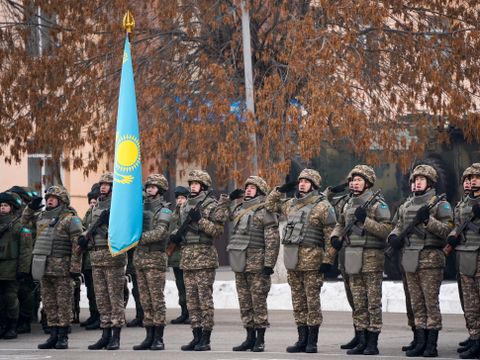 En gruppe menn i militære kamuflasjedrakter og hjelmer står på rekke bak et turkist og gult flagg. 