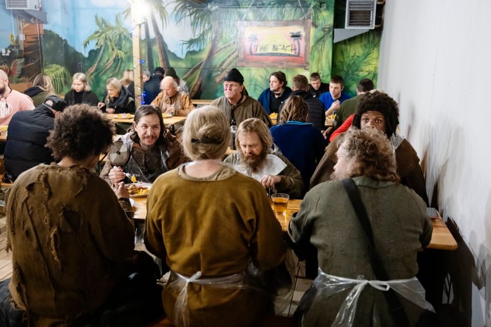 En gruppe mennesker i en spisesal ved flere bord, hvor halvparten har middelalderdrakter og halvparten er kledd i moderne klær.