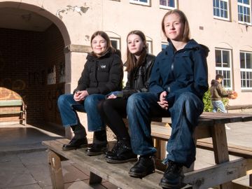 Tre jenter med mørke klær sitter ute på et bord, utenfor en skolebygning. 