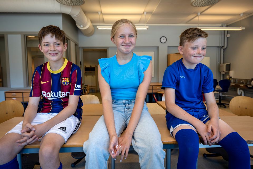 Tre barn i barneskolealder, to gutter og én jente, sitter på en pult inne i et klasserom og smiler.