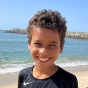 Nærbilde av en solbrun gutt med krøllete, brunt hår, som smiler bredt mens han står på en strand.