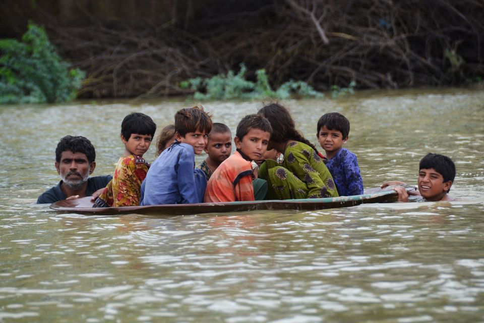 En voksen og et barn står i dypt, brun-grønt vann og holder en stor plate som det sitter åtte barn med bekymrede miner og fargerike klær oppå.
