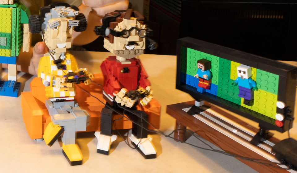 To gutter som sitter i en gul sofa og gamer på en liten tv-skjerm ved siden av bygget i lego. 