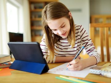 En jente i stripete genser sitter lent over et bord og skriver på et ark med et nettbrett foran seg