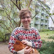 En dame med kort hår og ullgenser som står og smiler ute foran en boligblokk og hun holder en stor brun høne i armene. 