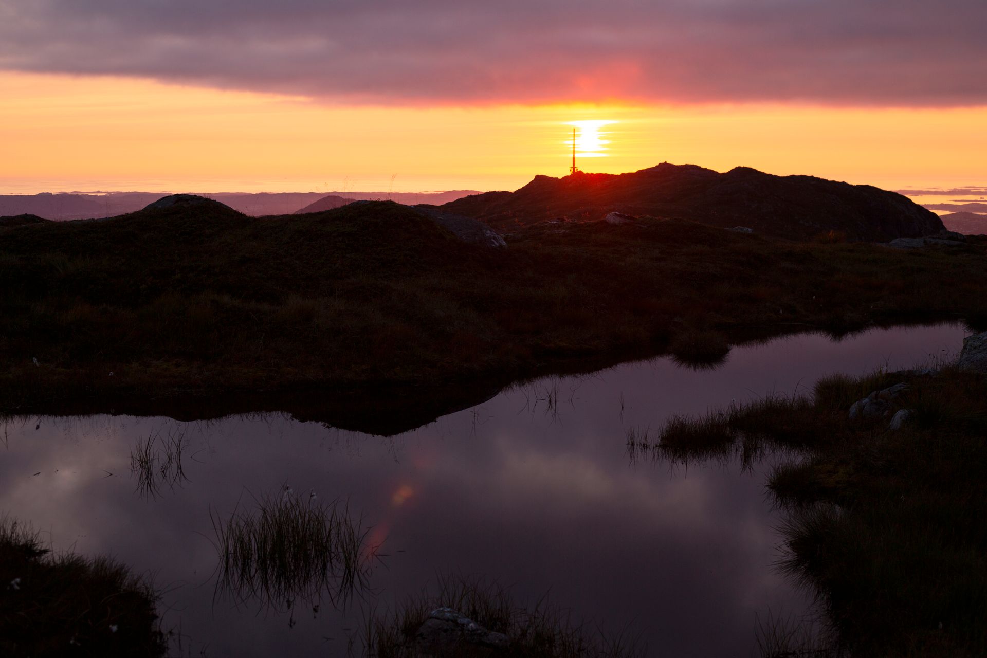 Landskapsfotografi av gresskledd vidde og et vann i rød-gul-lilla solnedgang, med utsikt mot radiomasten på toppen av Ulriken og havet i horisonten.