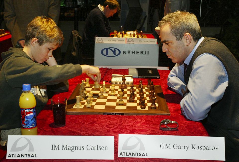 En ung gutt spiller sjakk mot en eldre mann på et rødt bord, og begge ser veldig konsentrerte ut. 