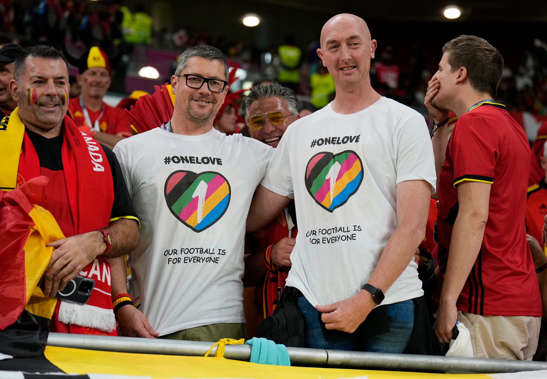 To menn står på en fotballtribune og smiler med hvite t-skjorter med regnbuefarger på.