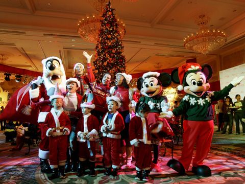 Personer utkledd som Mikke Mus og flere av vennene hans står ved et juletre og synger sammen med små barn i røde nissedrakter.