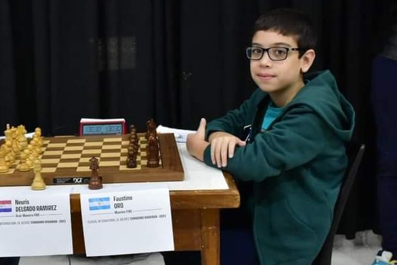 En ung gutt med briller sitter ved et lite bord med en tre-sjakkbrett og en stoppeklokke på.