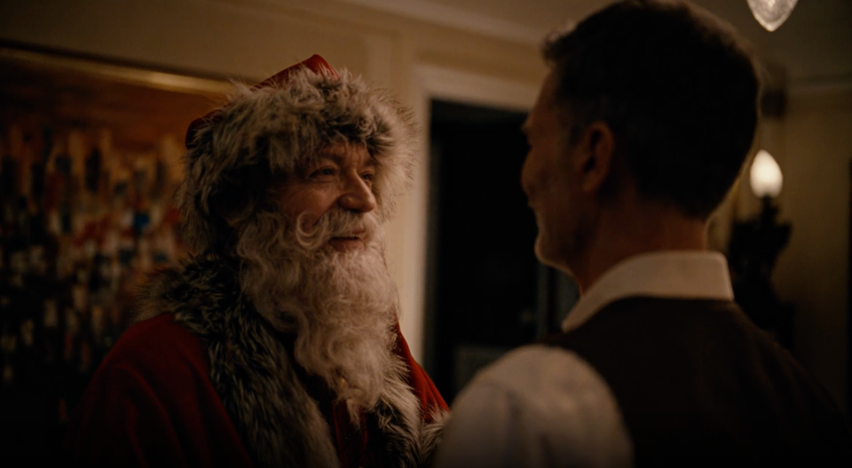 En mann med hvit skjorte og svart vest står med ryggen til fotografen og smiler mot ansiktet til en julenisse med rød lue og gråhvit brem, langt hvitt skjegg og snurrebart.