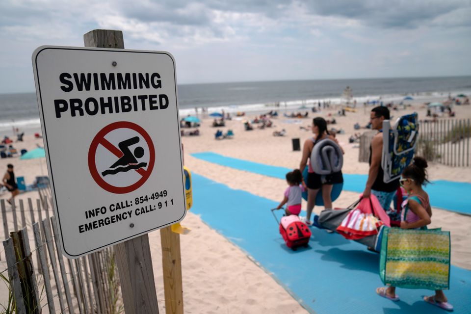 Flere personer går inn på en stor strand og går forbi et skilt som advarer mot å svømme.