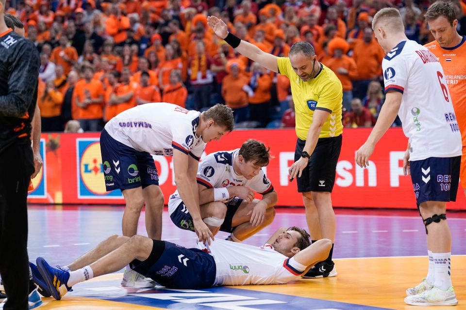 En mannlig håndballspiller ligger på gulvet og flere lagkamerater med hvite drakter står på huk rundt og ser om det går bra.