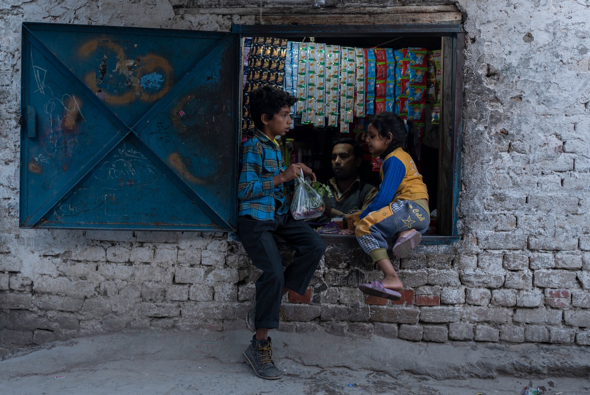 En gutt og jente sitter i et vindu i en liten kiosk. Gutten holder en pose med godteri som har akkurat har kjøpt. 