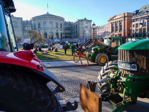 Flere traktorer i grønt og rødt står parkert foran Stortinget i Oslo på en dag med sol og blå himmel.