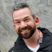 Nærbilde av en mann med mørkt skjegg og sort jakke som smiler mot fotografen.