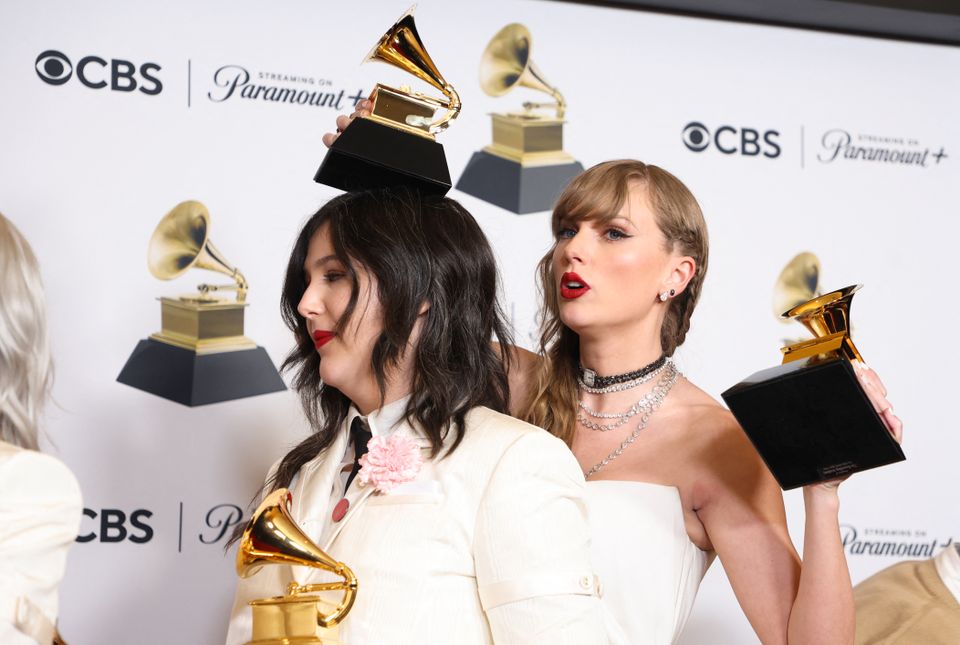 Taylor Swift, med langt, lyst hår og hvit kjole, holder flere pokaler, og den ene holder hun over hodet på en kvinne med langt, mørkt hår.