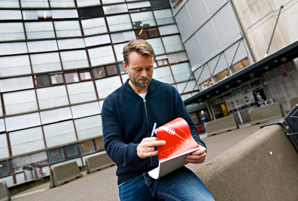En mann med mørk genser sitter på en stenblokk utenfor en stor bygning og kikker ned i den røde notisboken sin. 
