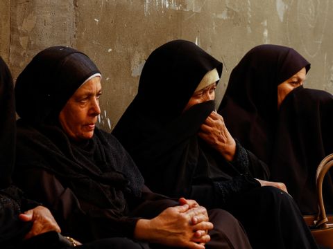 Tre kvinner med svarte hodeplagg og svarte, heldekkende kjoler sitter inntil en murvegg og gråter. 