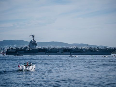 Et enormt skip med mange fly på, militærskipet Gerald Ford, ses på avstand i blått vann, med mange småbåter med tilskuere rundt.