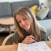 En jente med langt lysebrunt hår smiler og sitter ved et bord og tegner på et ark.