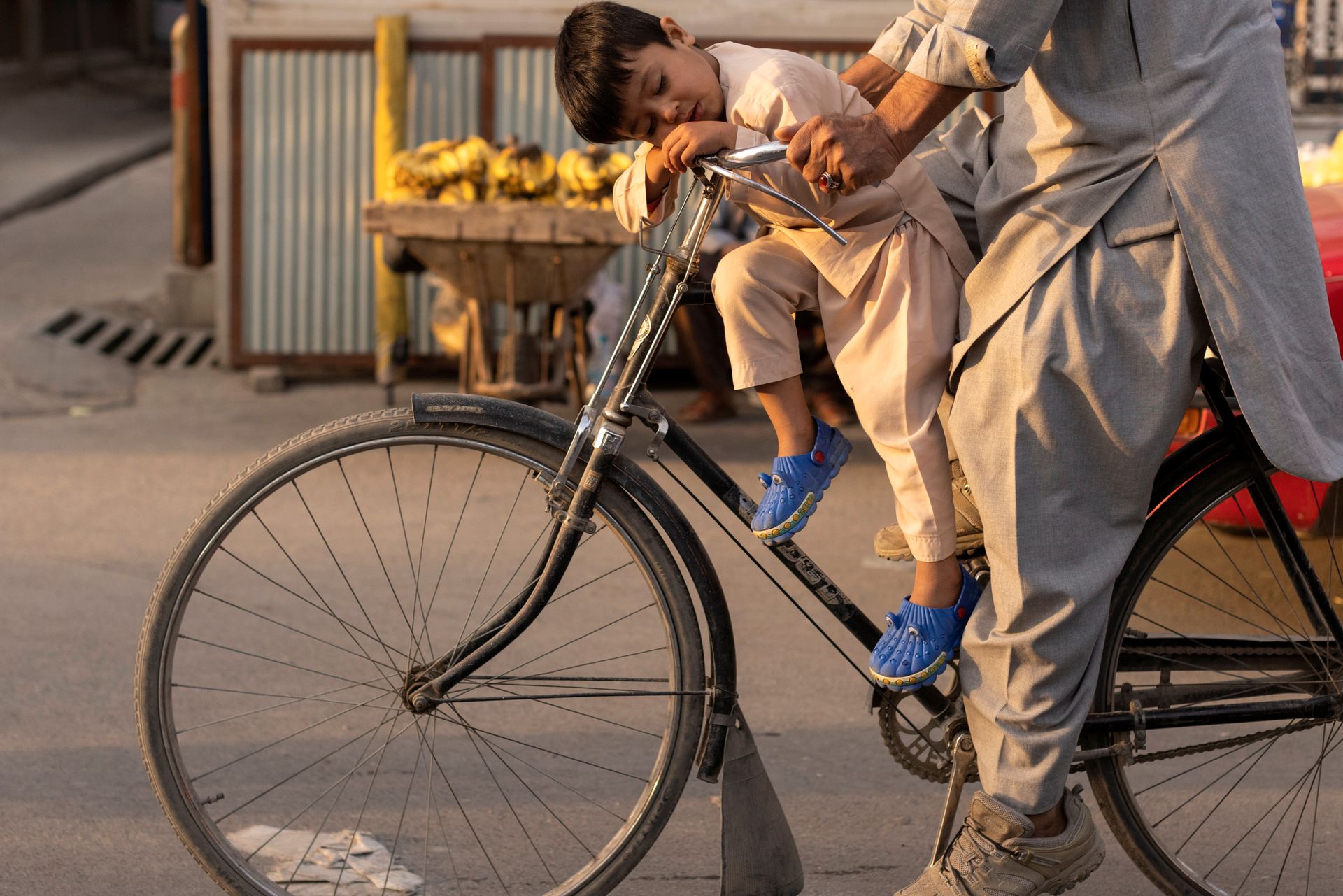 En gutt med svart hår, beige klær og blå sko sover på styret til en svart sykkel.