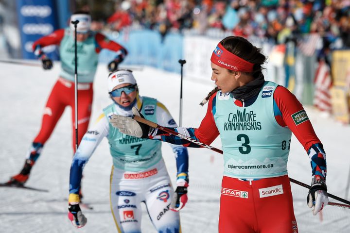 En kvinne med rød langrennsdrakt går på ski og har akkurat gitt et slengkyss og i bakgrunnen går flere konkurrenter.