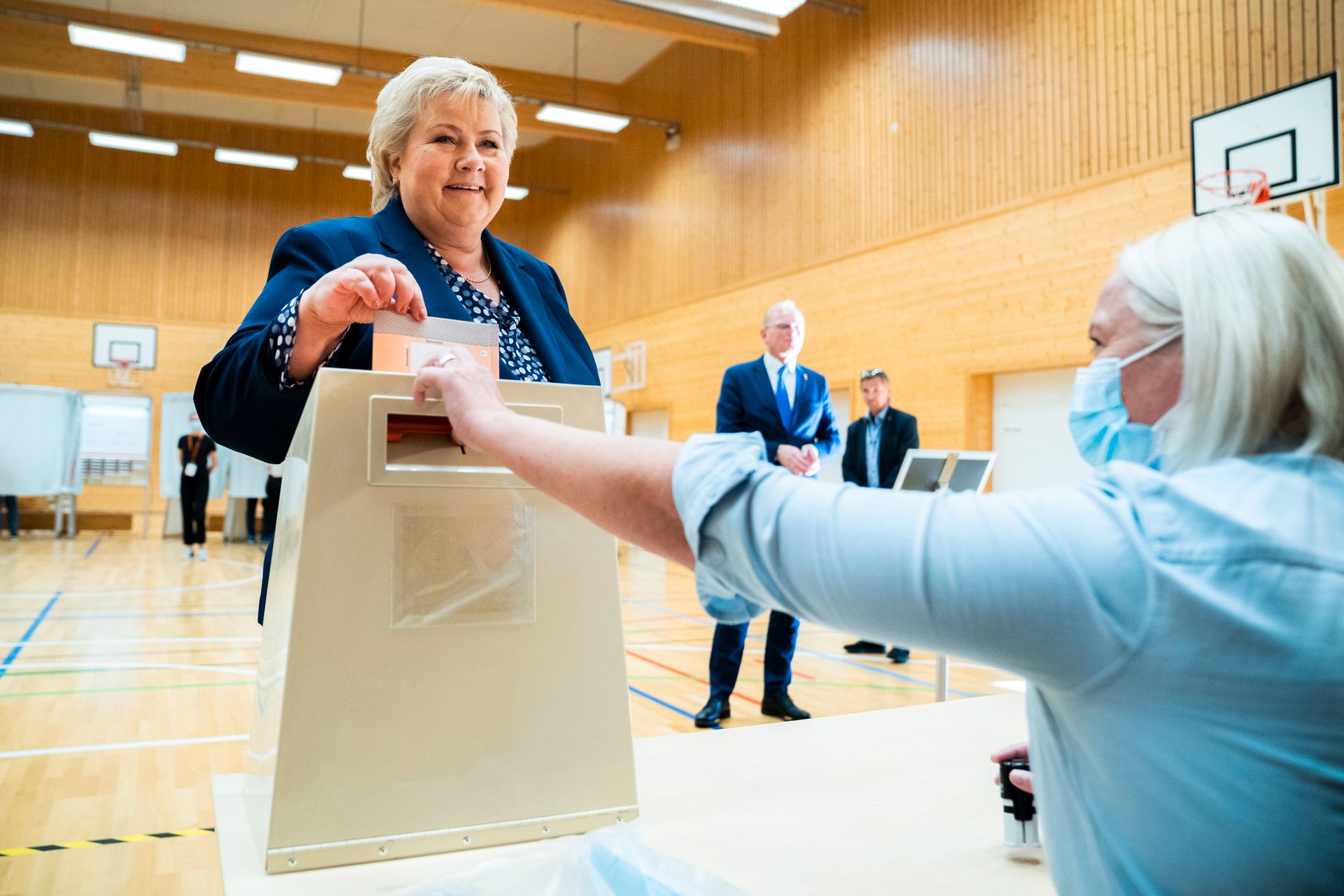 Erna Solberg smiler mens hun slipper sin stemmeseddel ned i valgurnen, som voktes av en blond kvinne med munnbind på.