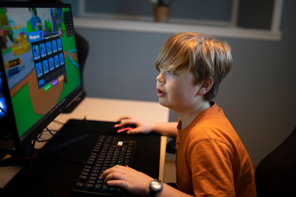 En gutt med lyst hår og oransje t-skjorte som sitter ved en PC med et spill på og trykker på tastaturet. 