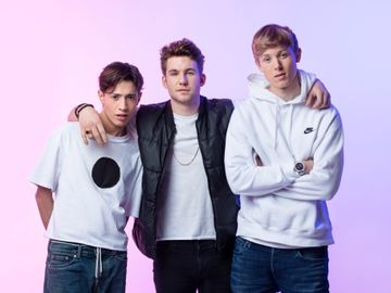 Tre unge gutter i hvite og sorte t-skjorter og hettegensere står tett med hånden rundt skulderen på hverandre foran en dus rosa bakgrunn. 