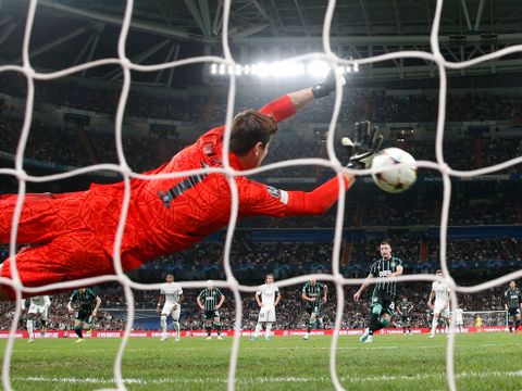 En mann i helt rød drakt kaster seg etter en fotball, sett gjennom nettet i et fotballmål.