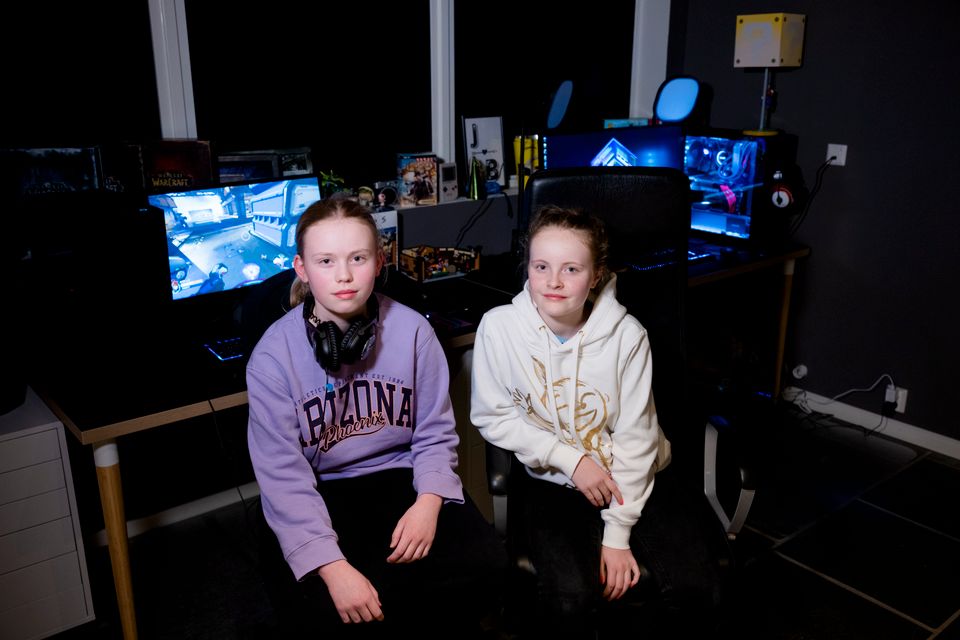 To jenter i lilla og hvit hettegensere sitter ved siden av hverandre i et mørkt rom med PC-skjermer som lyser i bakgrunnen. 