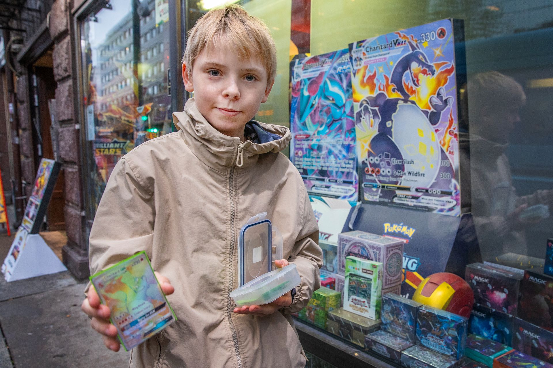En gutt med lyst hår som står utenfor en butikk med figurer i vinduet og han holder frem et kort med en drage på i den ene hånden og en plastboks i den andre. 