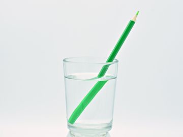 Bildet viser et vannglass med en grønn blyant i, som ser knekt ut, mot en hvit bakgrunn.
