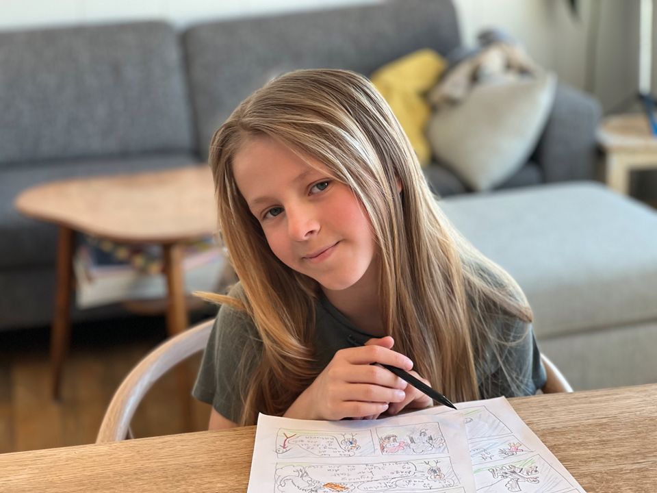 En jente med langt lysebrunt hår smiler og sitter ved et bord og tegner på et ark.